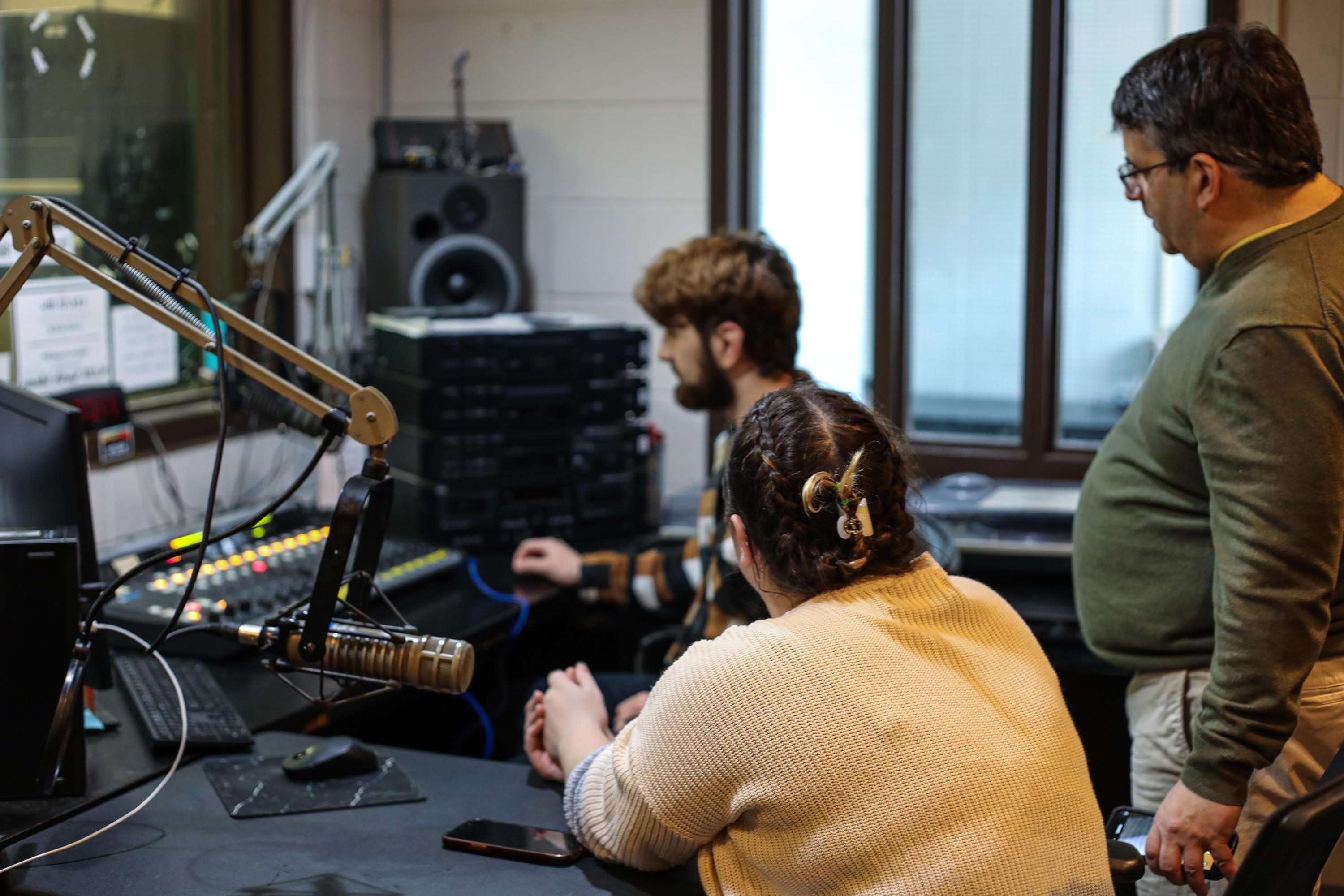 三个学生坐在广播演播室里，周围都是麦克风和开关板. 准备直播. 